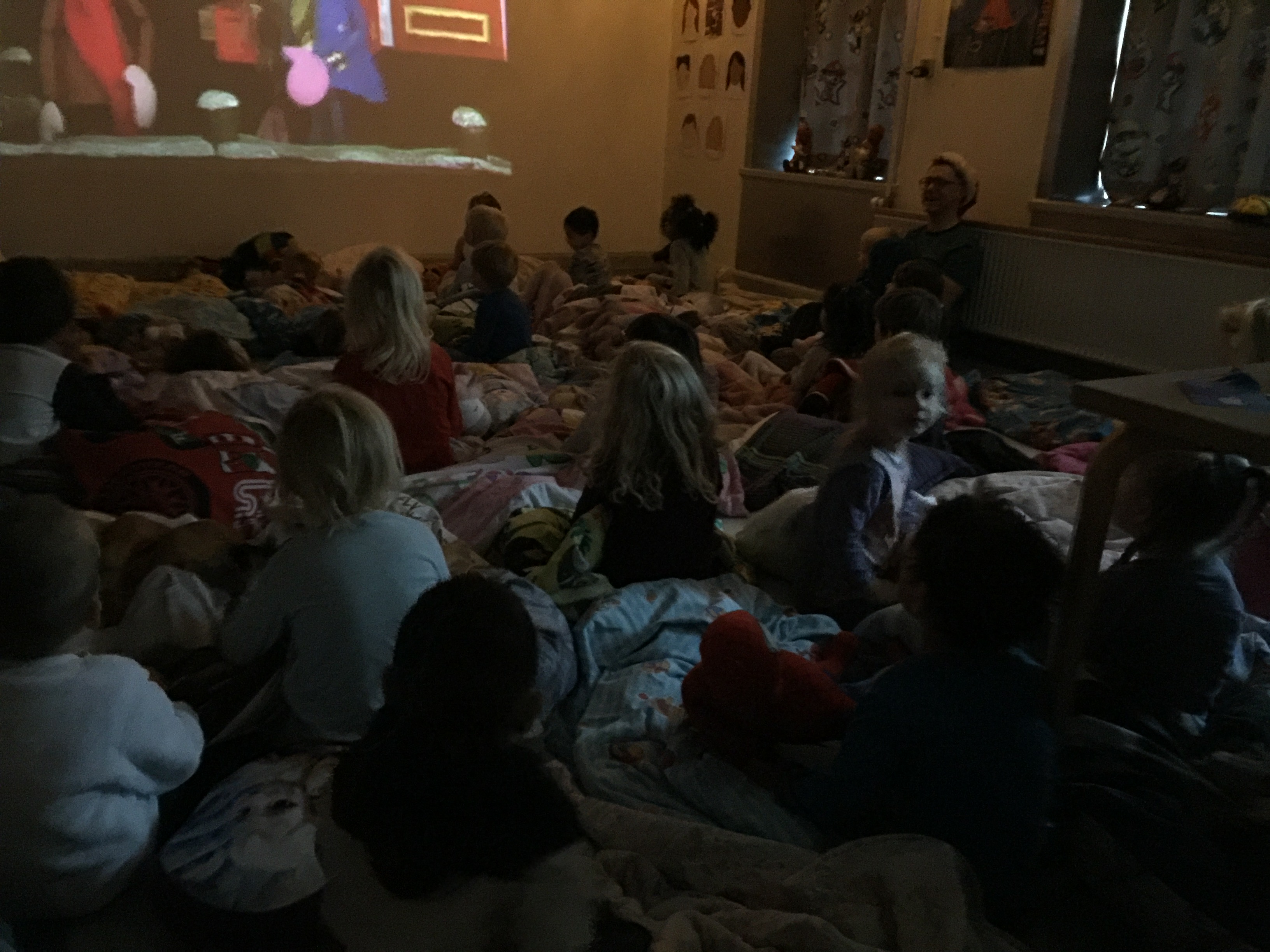 En masse børn sidder i deres dyner på gulvet og ser film på en stor skærm 